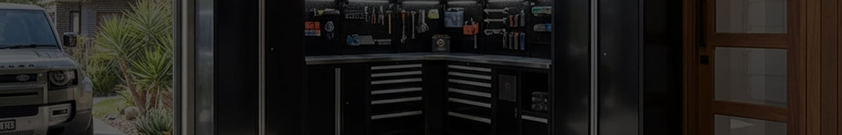 A dark, wide-angle view of a garage interior showing a black tool cabinet with drawers and shelves, alongside a white vehicle on the left and a wooden door on the right.